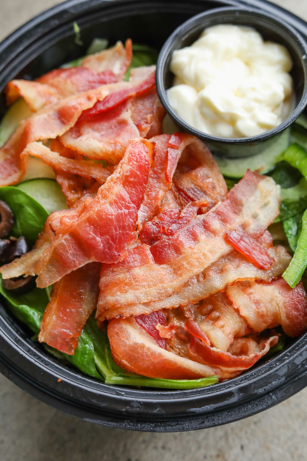 A black bowl filled with green vegetables, black olives, slices of bacon, and a side of mayonnaise.