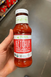 A hand holding a bottle of organic unsweetened ketchup.