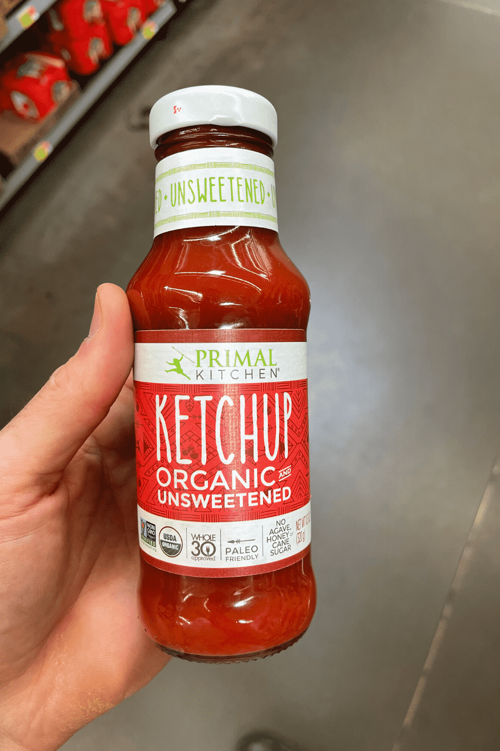 A hand holding a bottle of organic unsweetened ketchup.