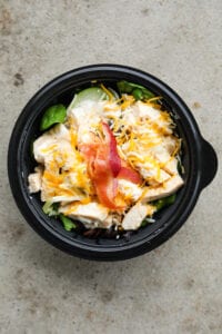 A black bowl filled with green vegetables, chopped chicken, ranch dressing, shredded cheese, and sliced bacon.