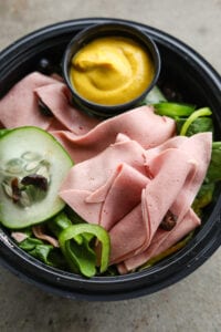 A black bowl filled with green vegetables, sliced bologna, and a cup of yellow mustard.