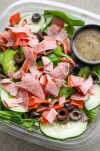 A plastic container filled with green vegetables, black olives, chopped up cold cuts, and a vinaigrette.