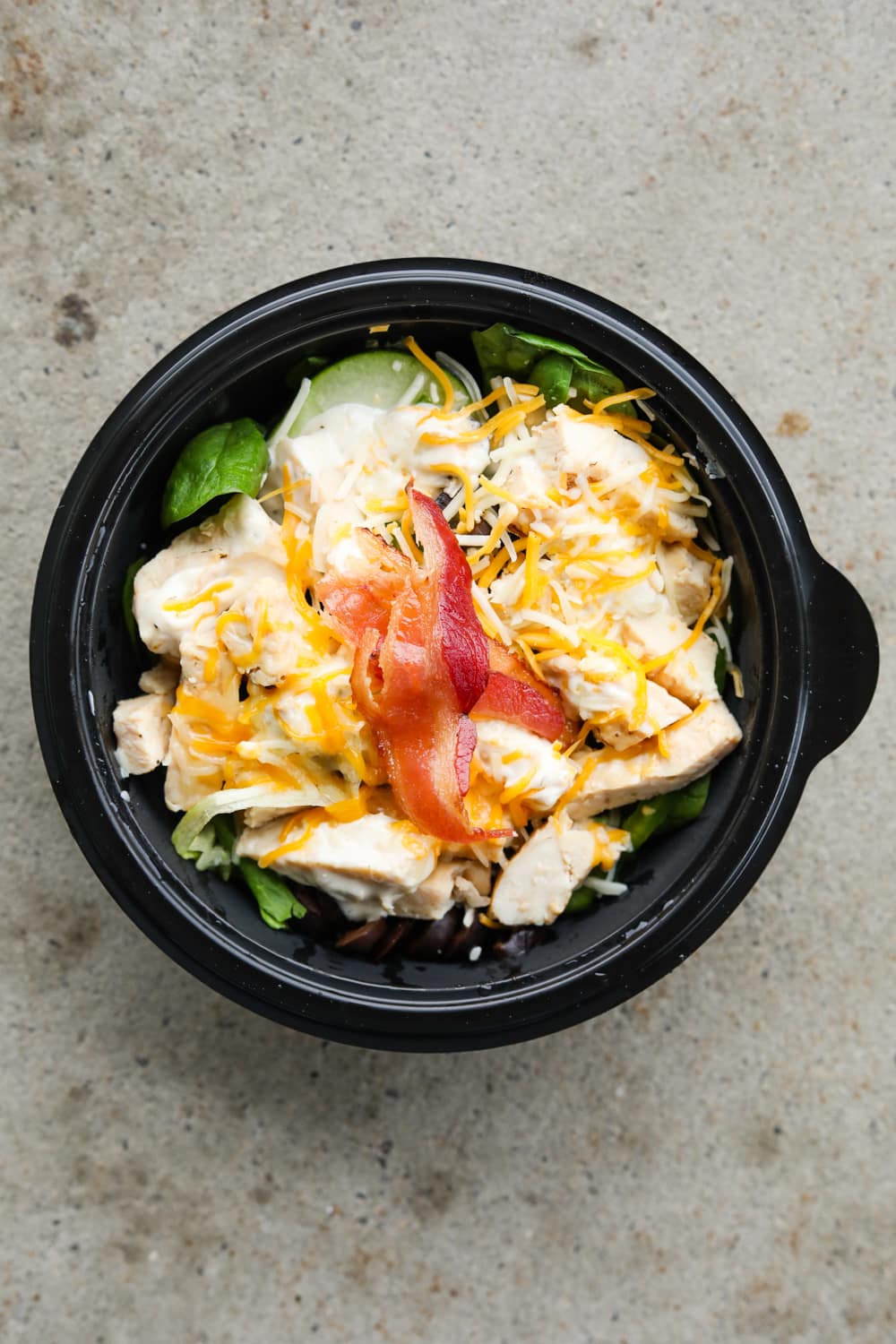 A black bowl filled with green vegetables, chopped chicken, ranch dressing, shredded cheese, and sliced bacon.