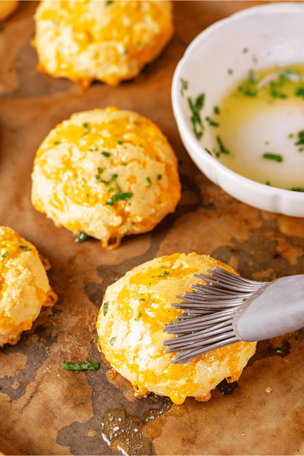 A few keto cheddar Bay biscuits on a piece of parchment paper. One of the biscuits is being brushed with a parsley garlic butter.