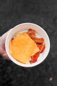 A hand holding a bowl filled with strips of bacon and eggs and American cheese and egg.