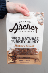 A hand holding a bag of country archer provisions hundred percent natural turkey jerky hickory smoke flavored.