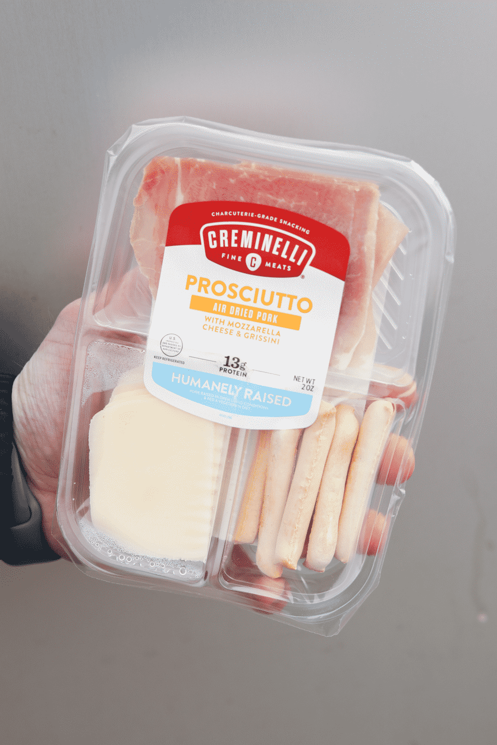 A hand holding a container of prosciutto, cheese and grissini.