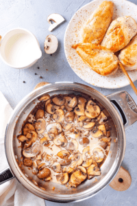 A pan filled with mushrooms and a creamy marsala sauce. Behind the pan is a white plate with four pieces of chicken on it.