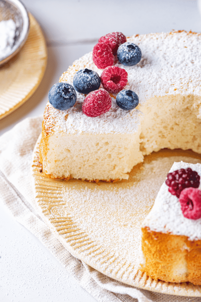 An angel food cake on a plate with a piece cut out of the front of the cake. There are blueberries and raspberries along with powdered sugar on top of the cake.