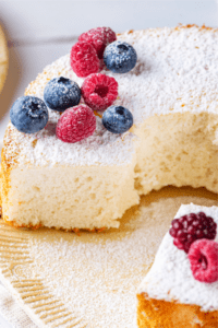 An angel food cake on a plate with a piece cut out of the front of the cake. There are blueberries and raspberries along with powdered sugar on top of the cake.