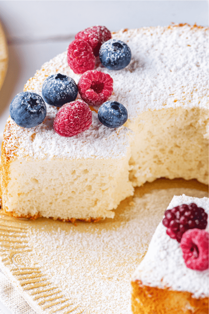 An angel food cake on a plate with a piece cut out of the front of the cake. There are blueberries and raspberries along with powdered sugar on top of the cake.