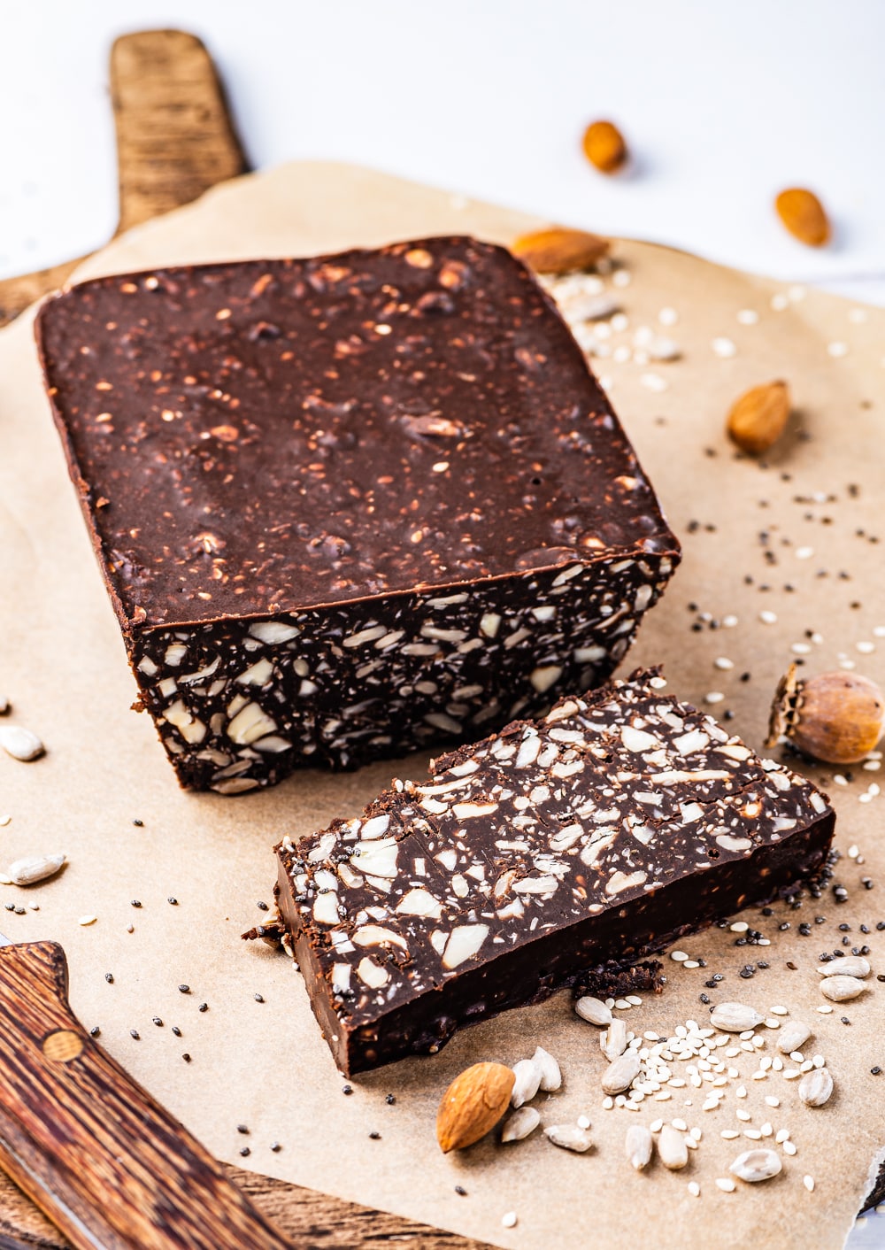 A block of chocolate filled with nuts on a cutting board. There is a slice of chocolate cut off from the block, and set in front of it.