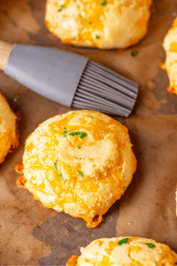 A keto cheddar Bay biscuits on a piece of parchment paper. There is a brush right behind the biscuit.