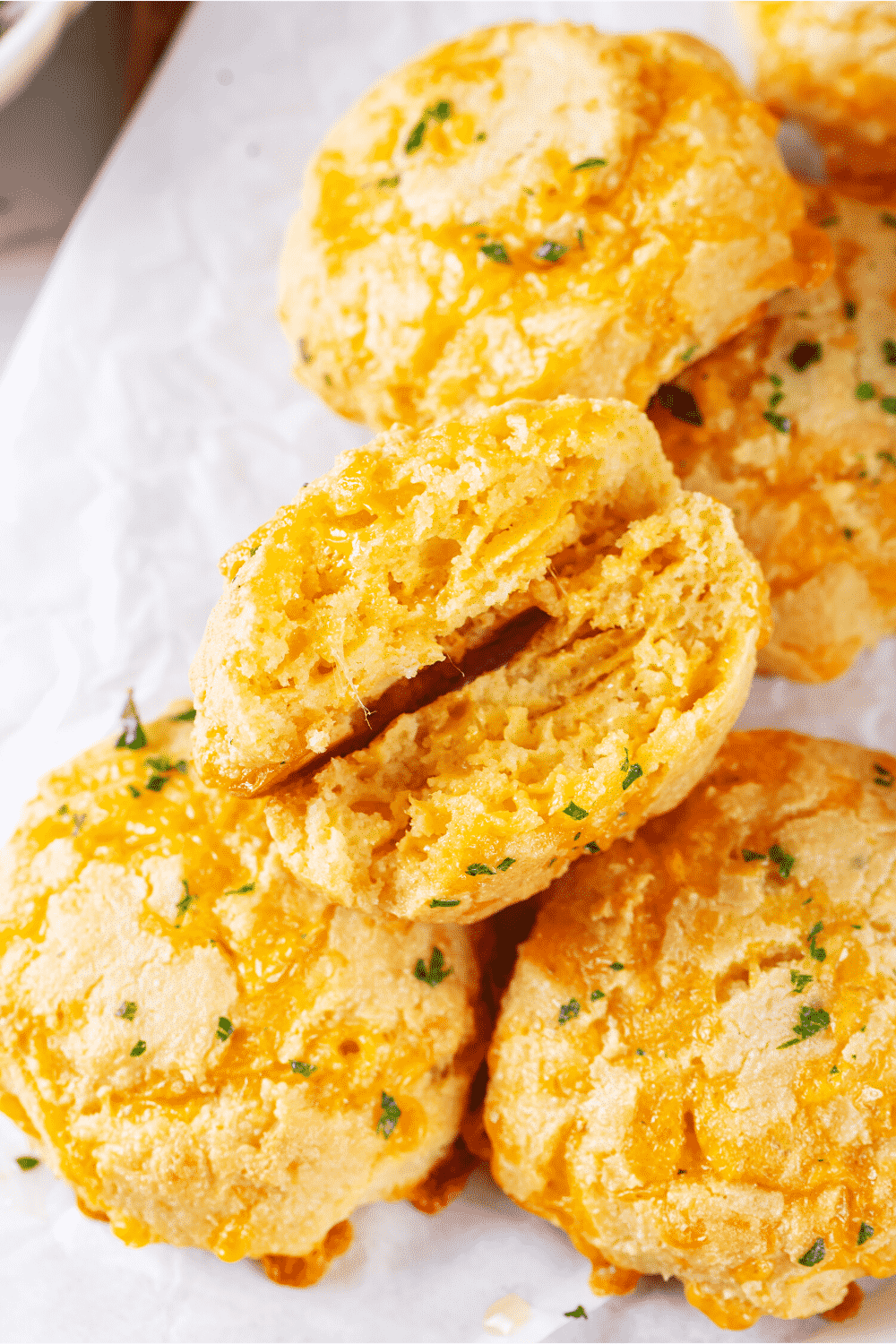A bunch of keto cheddar Bay biscuits on a piece of white parchment paper. One biscuit is broken in half with the insides of the biscuit facing up.