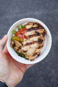 A hand holding a cup filled with a piece of grilled chicken lettuce tomatoes and pickles.