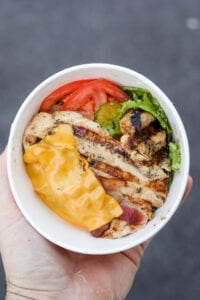 A hand holding a cup filled with a piece of grilled chicken, bacon, a slice of American cheese, lettuce, pickles, and tomato.