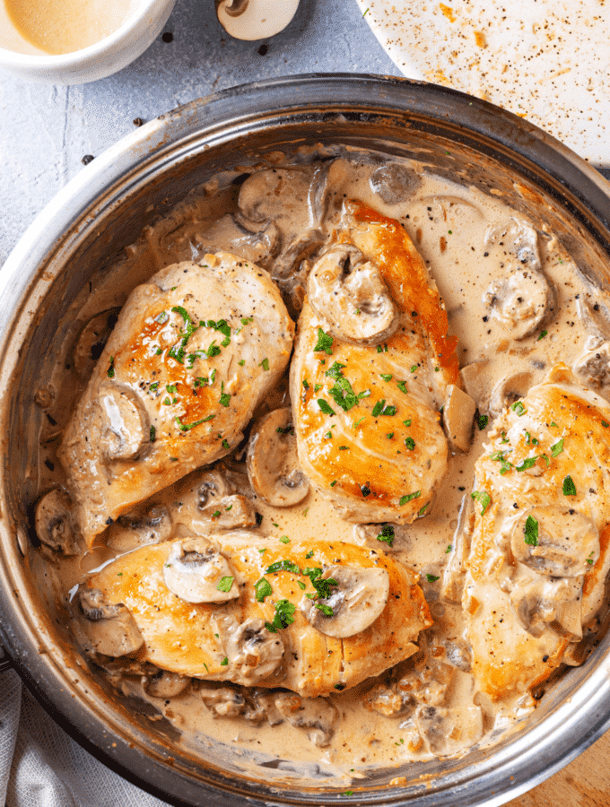 A pan filled with four pieces of chicken marsala. The pan is on a wooden cutting board on a blue counter.