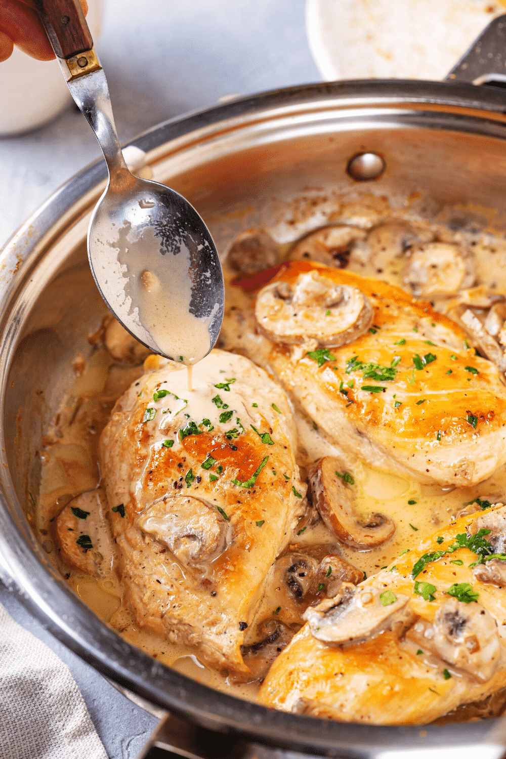 A pan with three pieces of chicken marsala in it. A spoon is drizzling marsala sauce over one piece of chicken.