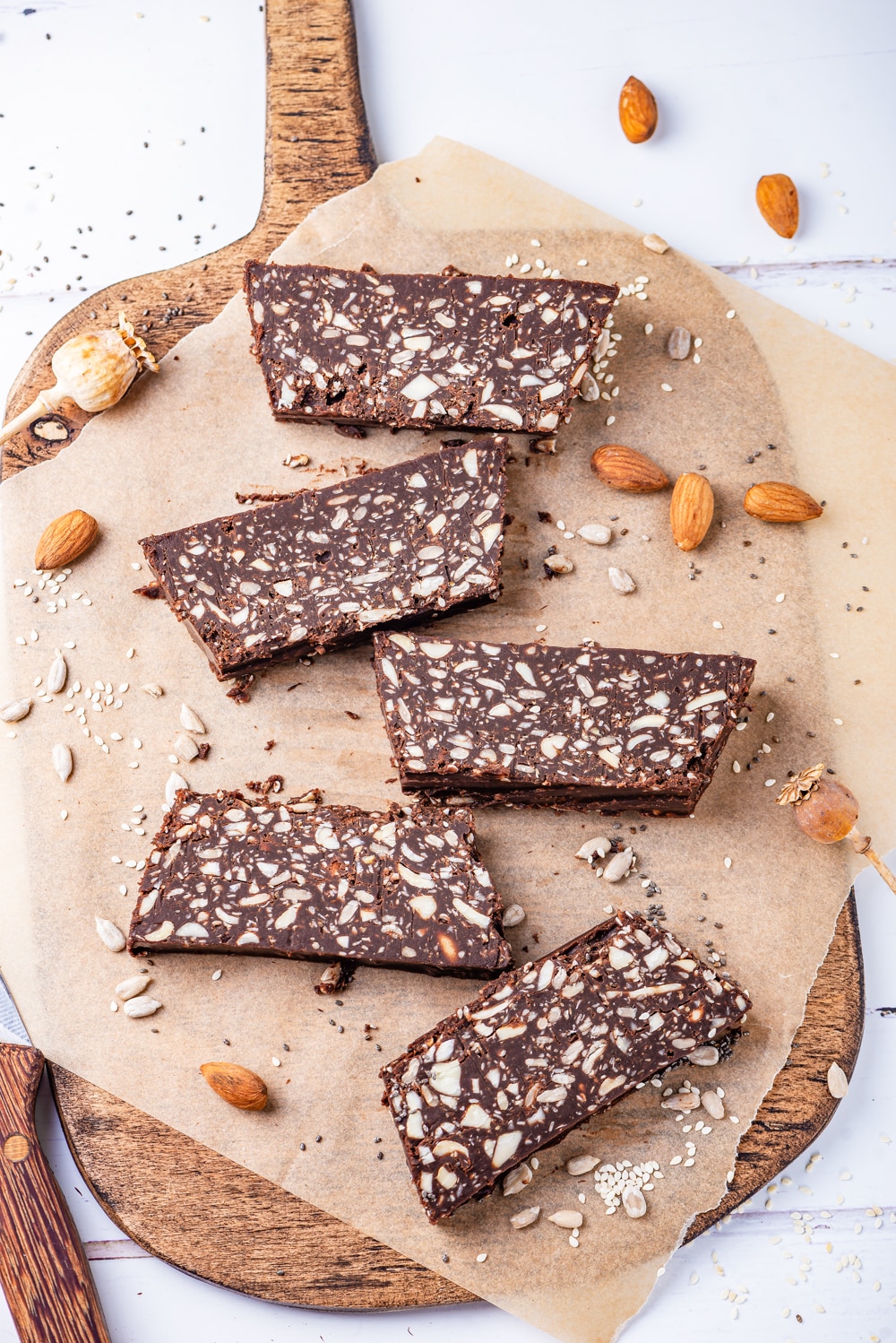 Five nut and seed filled chocolate bars on a brown sheet of parchment paper that's set on a wooden cutting board.