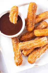A bunch of churros on a piece of white parchment paper. There is a cup of chocolate sauce to the left of the churros and one churro l is submerged in the chocolate sauce.