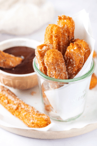 Glass cup filled with six churros. The glass cup is on a piece of white parchment paper on a plate and there is one churro next to the cup on the parchment paper and a cup of chocolate sauce behind it.
