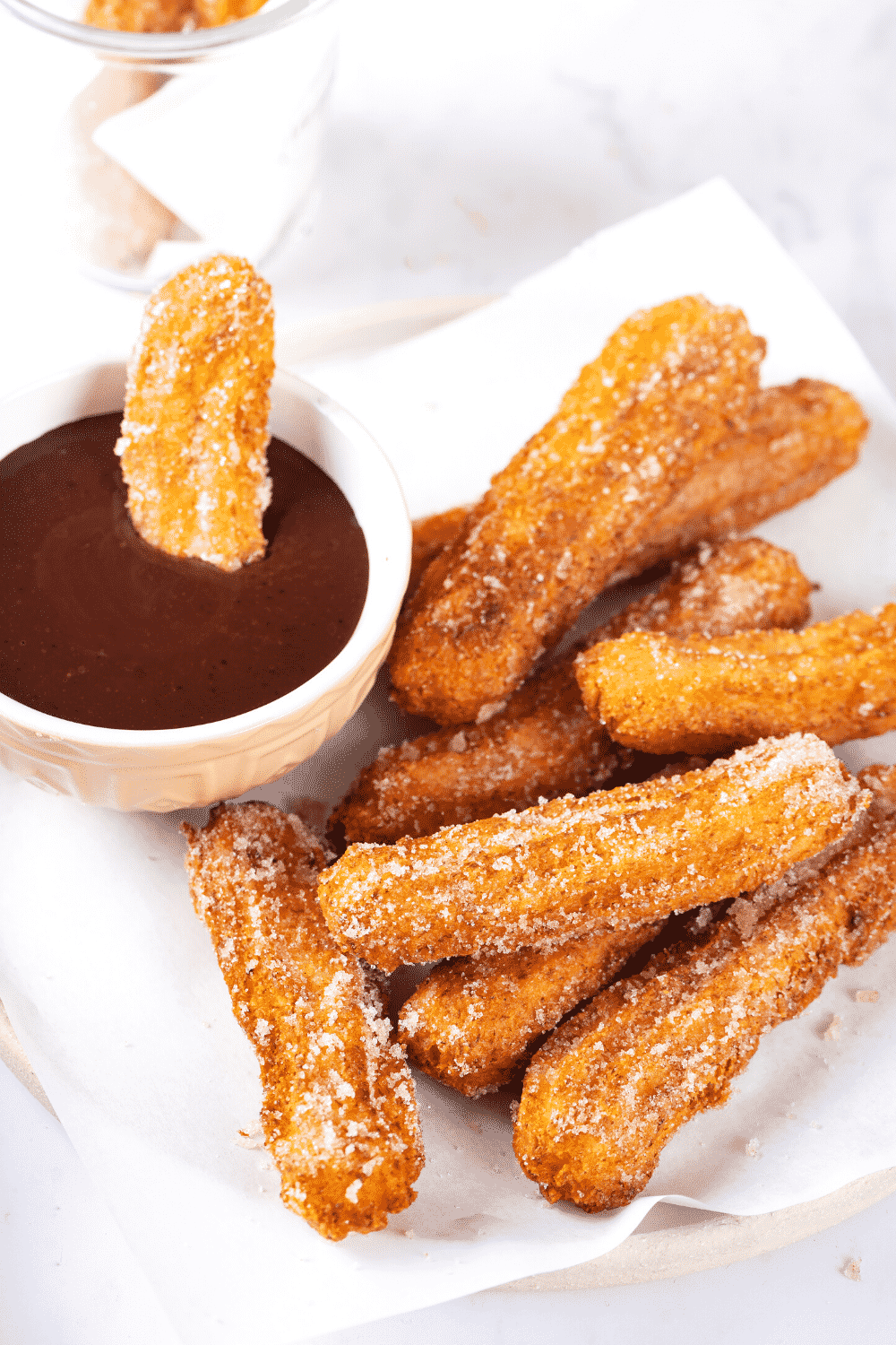 A bunch of churros and a piece of white parchment paper on a plate. There is a small cup of chocolate sauce to the left on the churros on the plate.