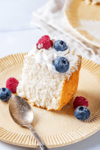 A piece of angel food cake on a plate with a raspberry and blueberry in front of and behind the piece of cake.