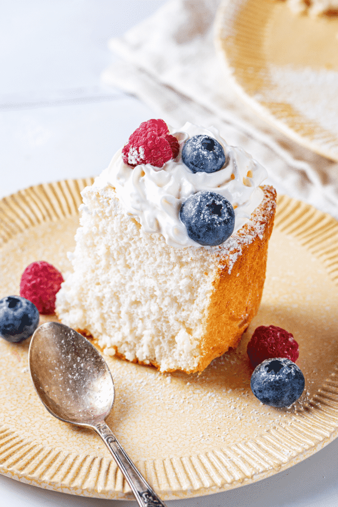 A piece of angel food cake on a plate with a raspberry and blueberry in front of and behind the piece of cake.