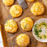 Southern keto cheddar Bay biscuits on a piece of parchment paper on a baking sheet. A bowl with a little parsley garlic butter is on the parchment paper along with a brush towards the back of the parchment paper.