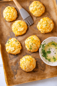 Southern keto cheddar Bay biscuits on a piece of parchment paper on a baking sheet. A bowl with a little parsley garlic butter is on the parchment paper along with a brush towards the back of the parchment paper.