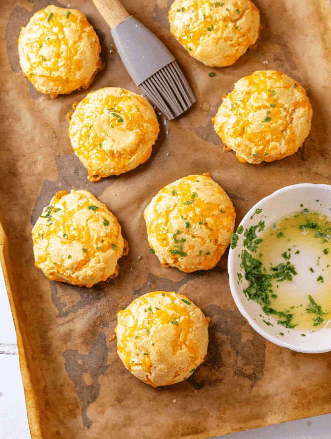 Southern keto cheddar Bay biscuits on a piece of parchment paper on a baking sheet. A bowl with a little parsley garlic butter is on the parchment paper along with a brush towards the back of the parchment paper.