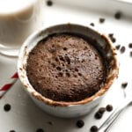 A chocolate cake in a white mug. There's a glass of milk, a straw, and a fork next to the mug.