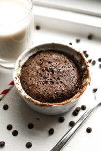 A chocolate cake in a white mug. There's a glass of milk, a straw, and a fork next to the mug.