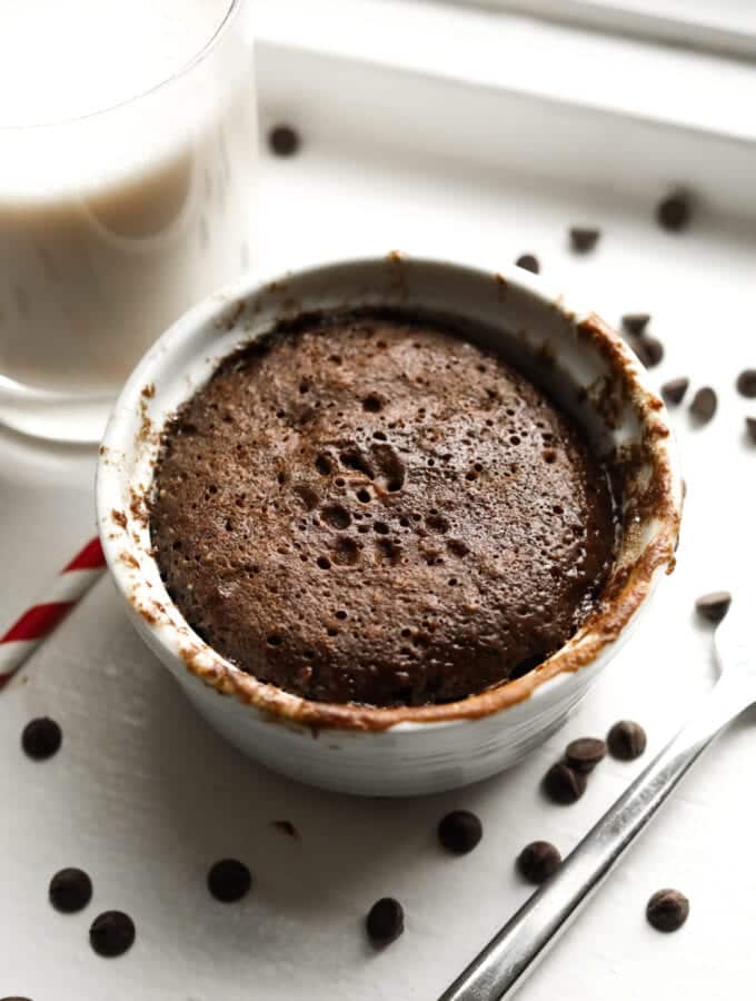 A chocolate cake in a white mug. There's a glass of milk, a straw, and a fork next to the mug.