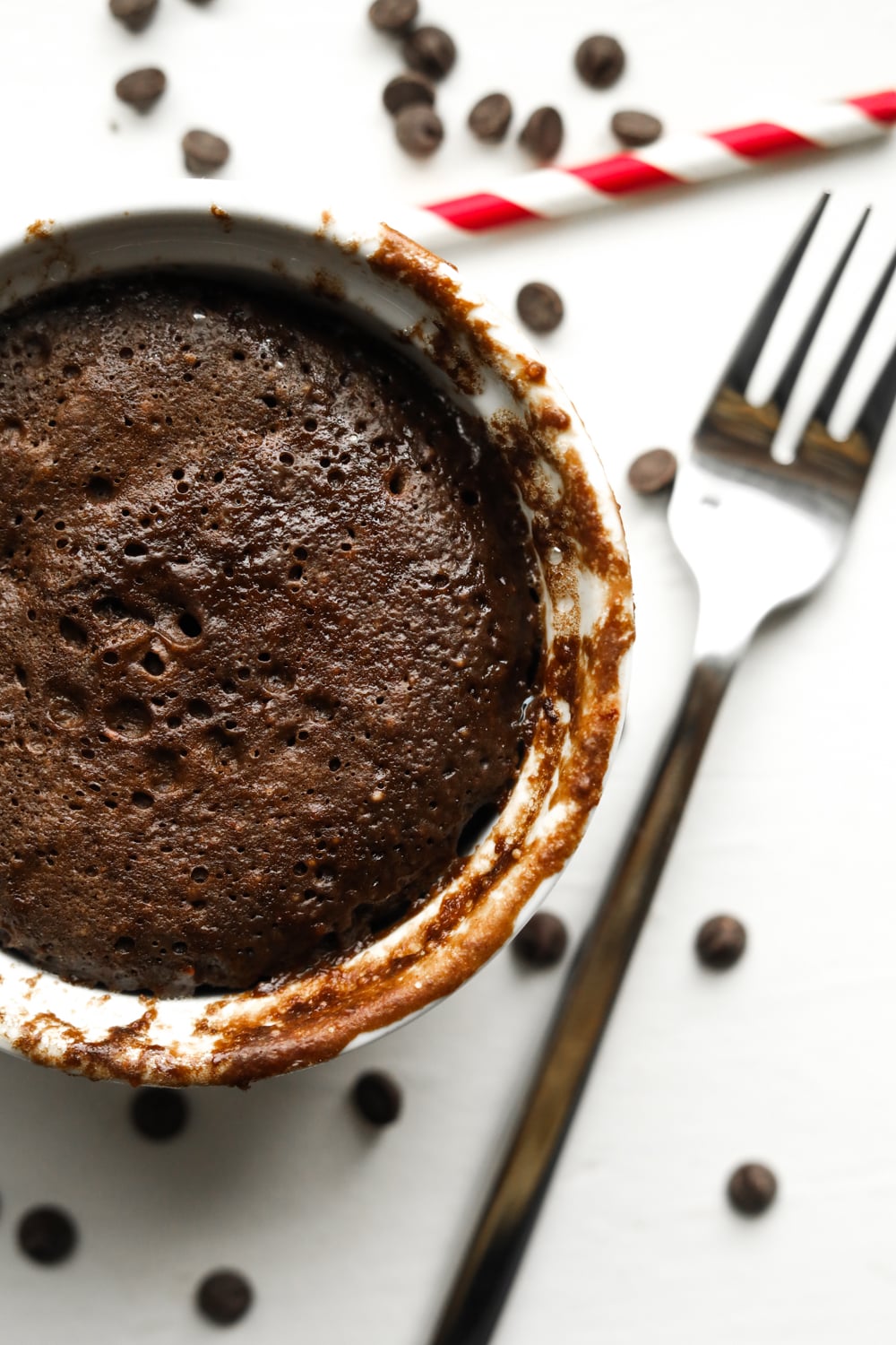 A chocolate cake in a white mug. There's a fork, chocolate chips, and a red & white straw next to the mug.