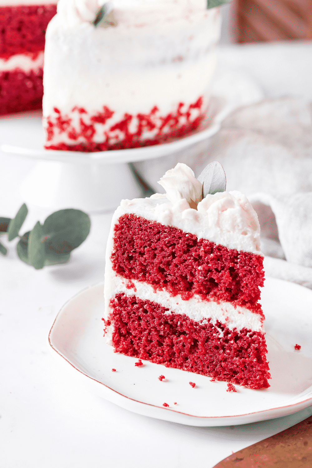 A white plate with a piece of red velvet cake on it. The rest of the red velvet cake is behind the piece on a white serving tray.