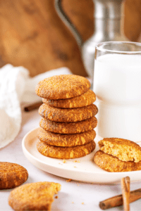 A stack of six snickerdoodle cookies on a white plate. There is a whole snickerdoodle cookie and half of one on top of it to the right of the stack. Behind that is a glass of milk.