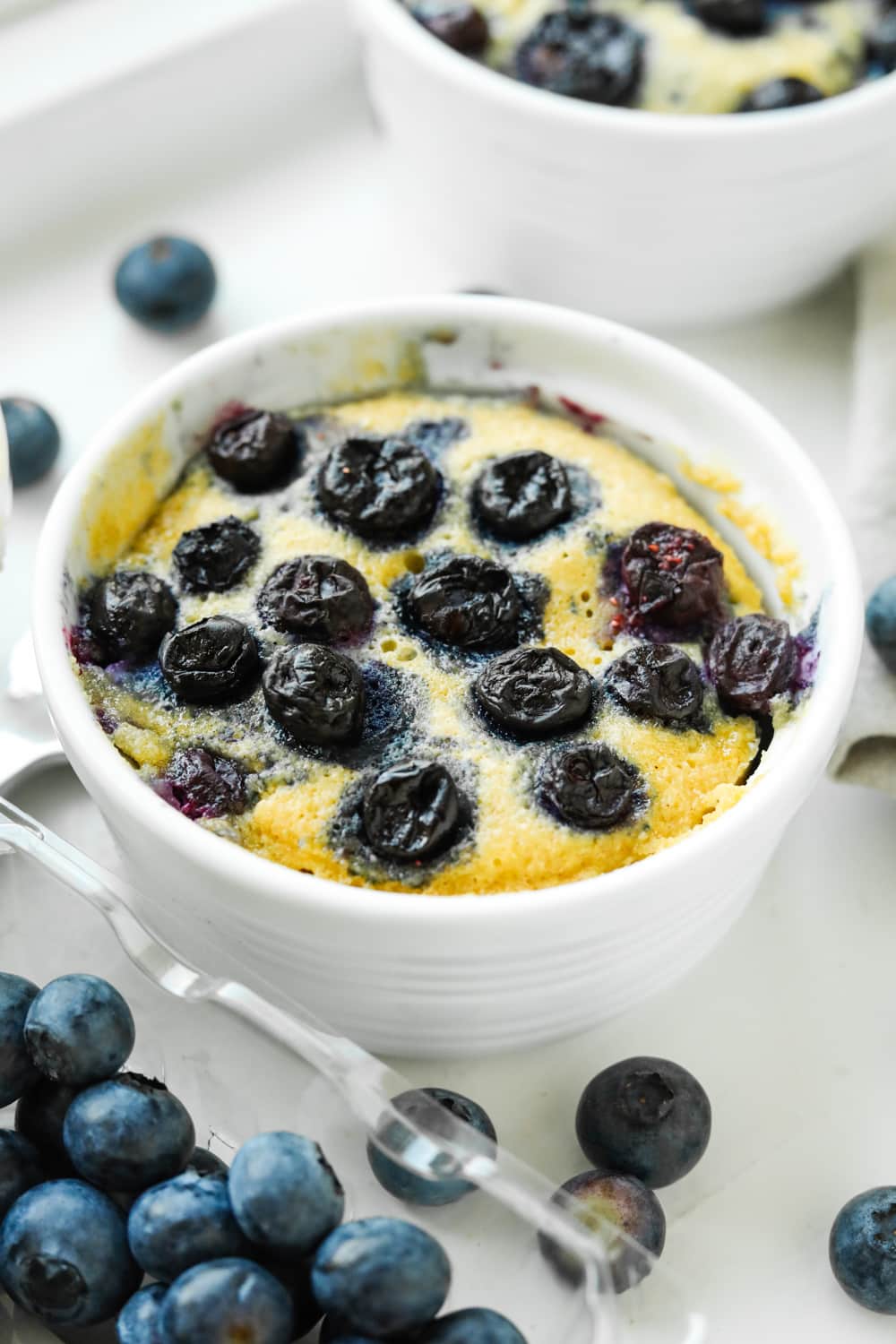 A blueberry muffin in a white mug with a container of blueberries in front of it, and another muffin place behind it.