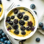 A blueberry muffin in a white mug. There are loose blueberries all around the mug.