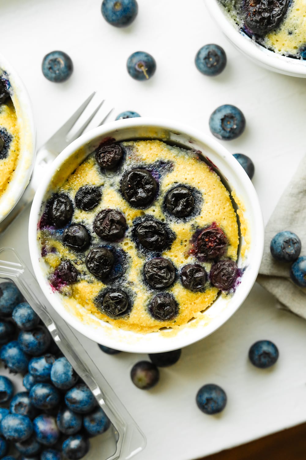 A blueberry muffin in a white mug. There are loose blueberries all around the mug.