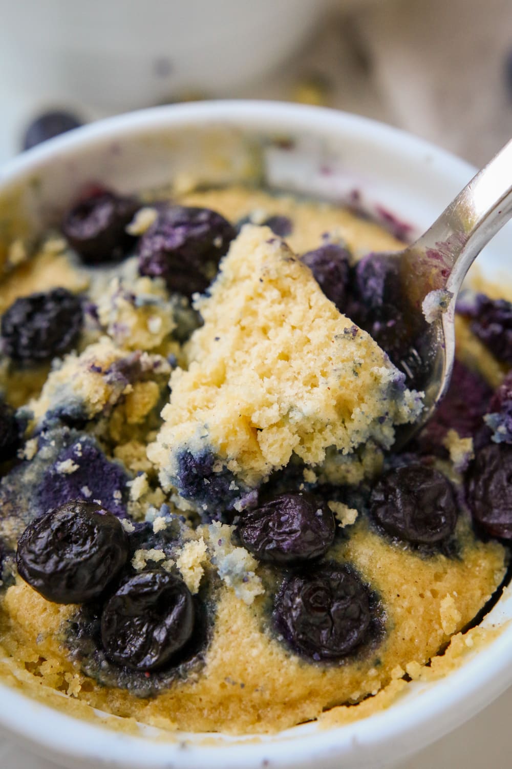 A fork holding a piece of a blueberry muffin. The for is placed on top of the muffin, and the muffin is inside a white mug.