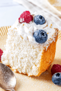 A piece of angel food cake on a plate. The piece of angel food cake has whipped cream and two blueberries and one raspberry on top.