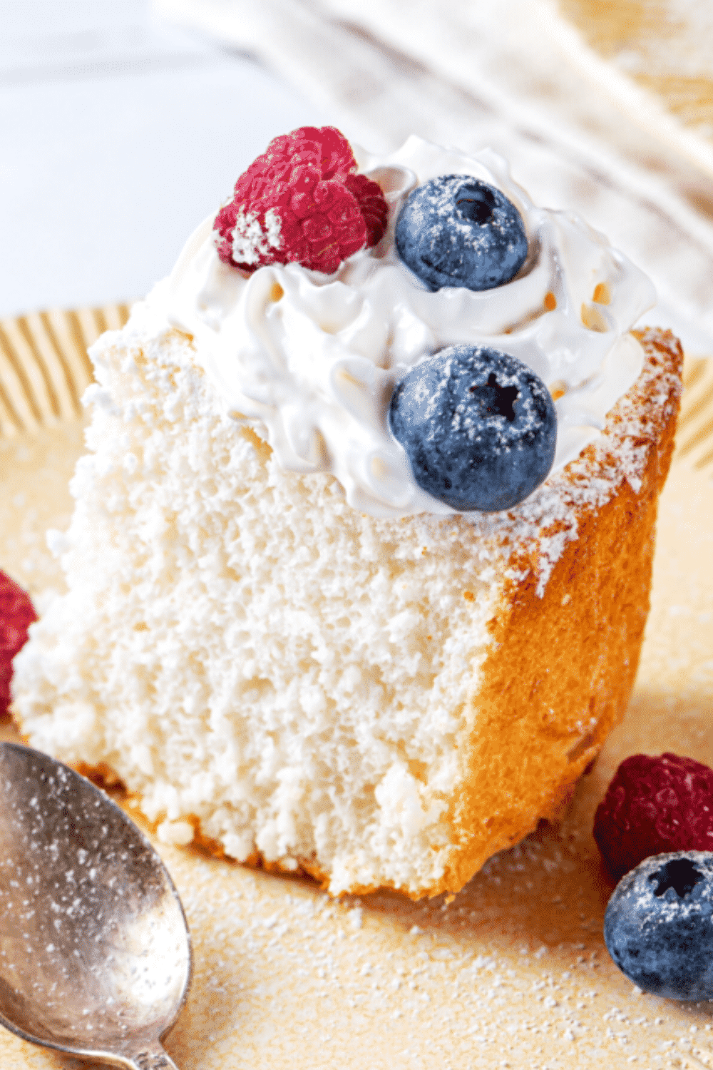 A piece of angel food cake on a plate. The piece of angel food cake has whipped cream and two blueberries and one raspberry on top.