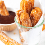 Six churros in a glass cup with white parchment paper raft behind them. The glass cup is on a piece of white parchment paper on a plate and there is one churro on the plate with a cup of chocolate sauce behind it.