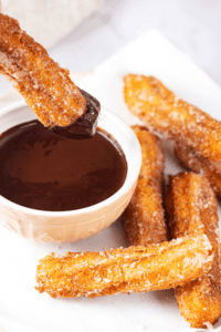 A couple of churros and a piece of white parchment paper. There is a cup of chocolate sauce to the left of the churros and one churro is dipped in the chocolate sauce.