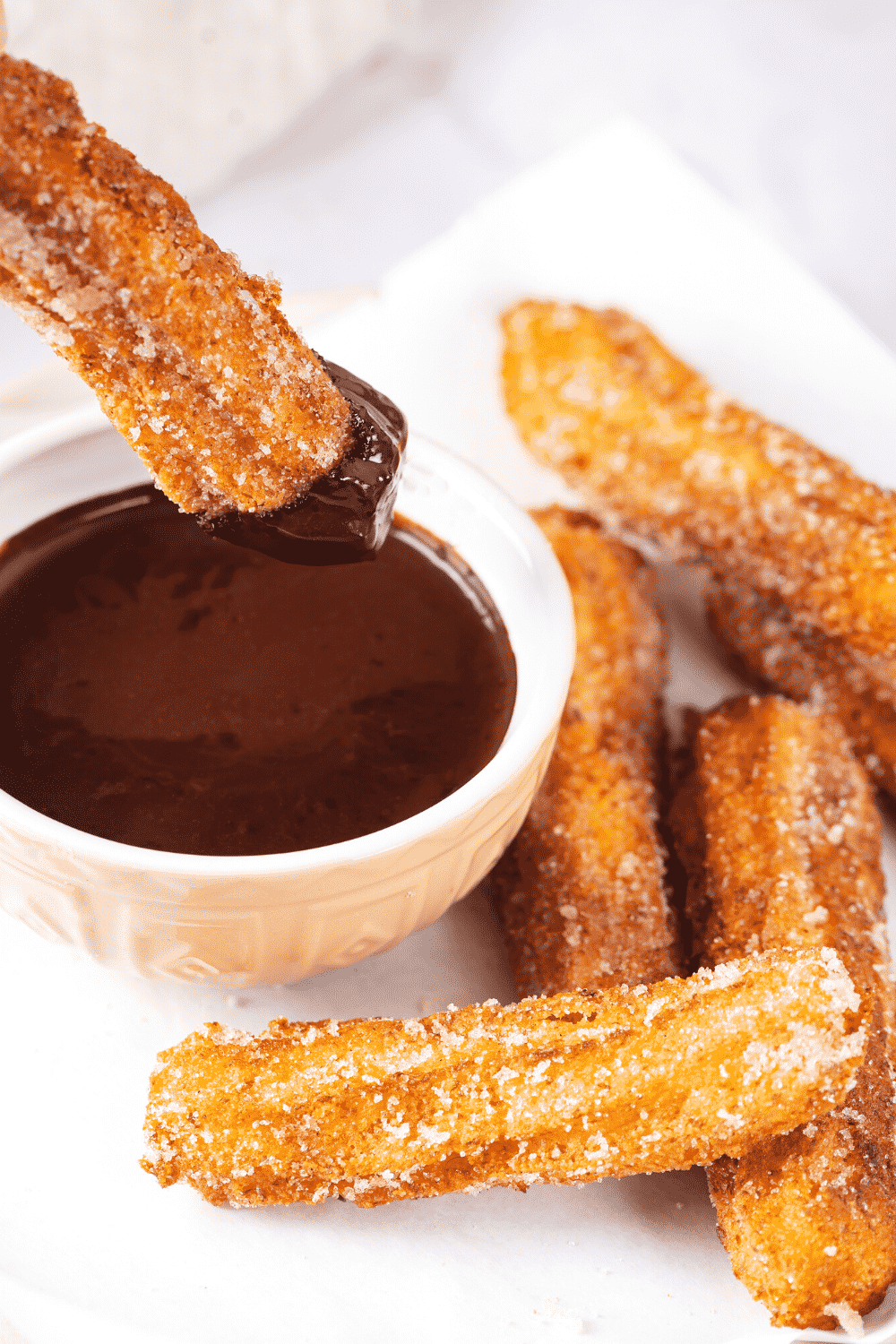 A couple of churros and a piece of white parchment paper. There is a cup of chocolate sauce to the left of the churros and one churro is dipped in the chocolate sauce.