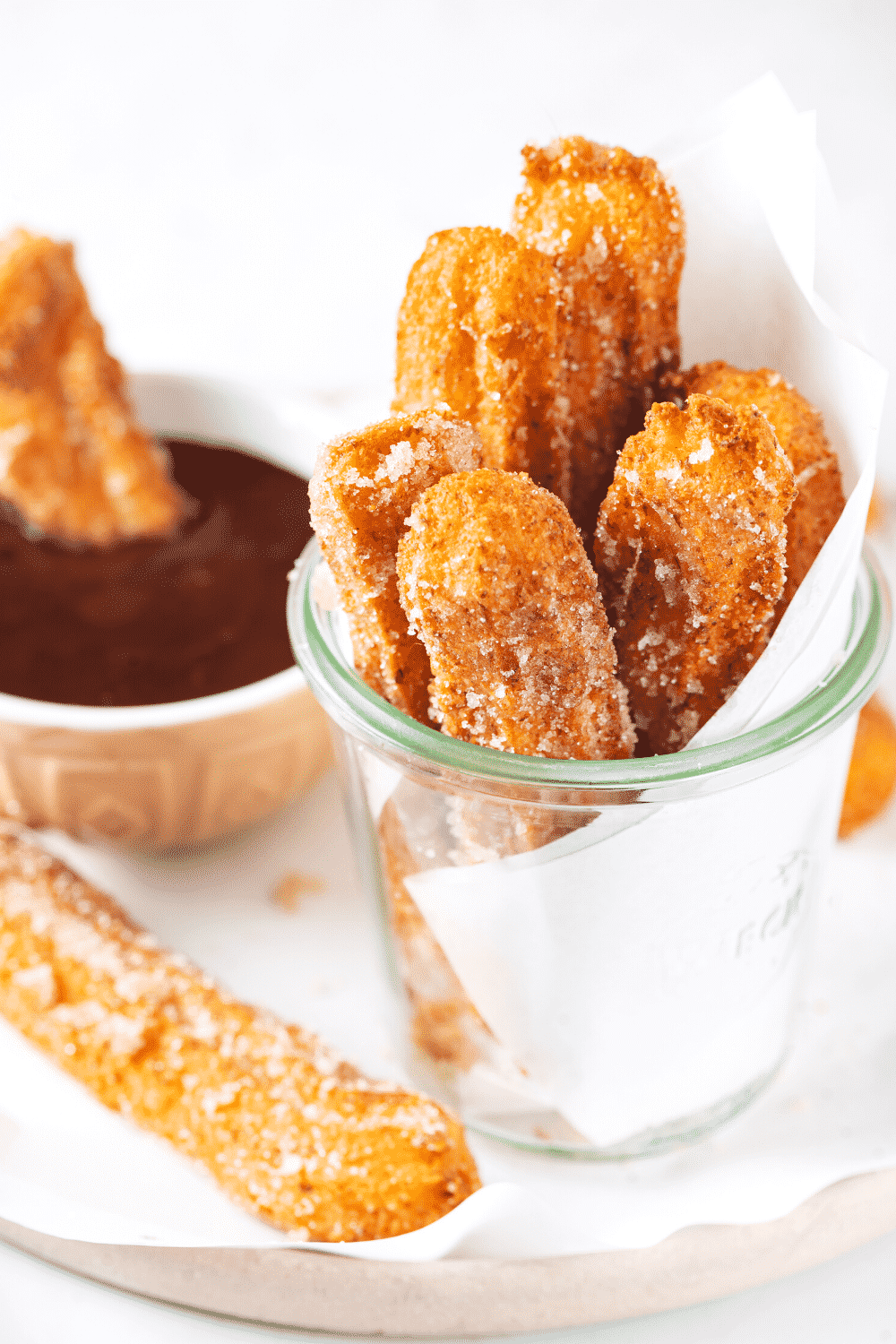 Six churros in a glass cup with white parchment paper raft behind them. The glass cup is on a piece of white parchment paper on a plate and there is one churro on the plate with a cup of chocolate sauce behind it.
