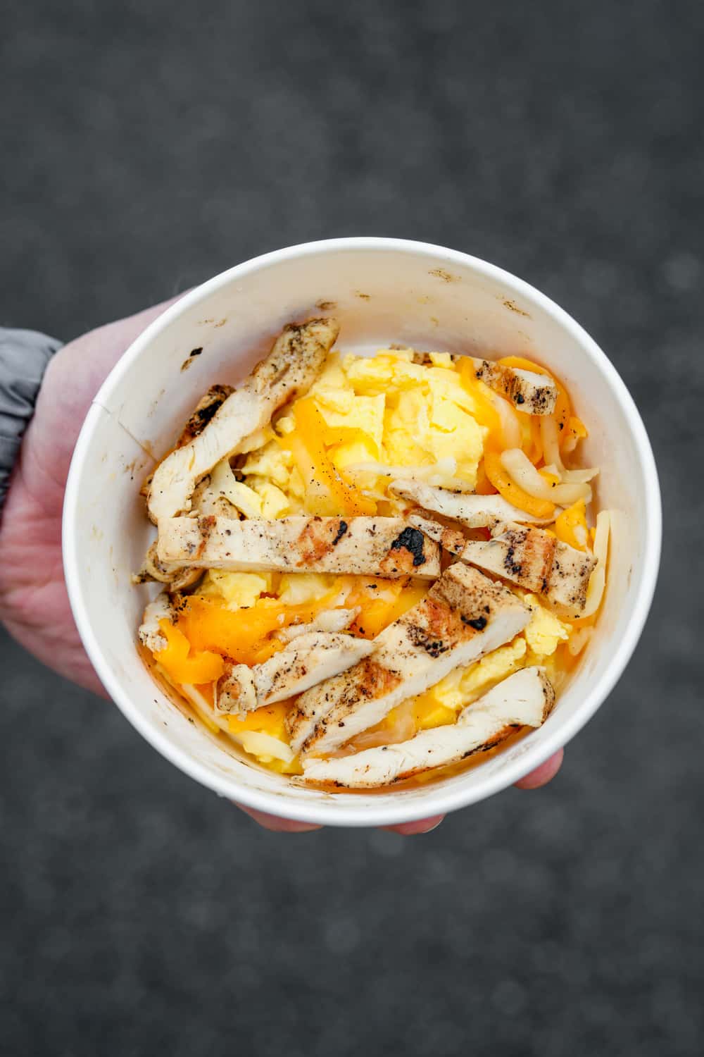 A hand holding a bowl filled with scrambled eggs Monterey and cheddar cheese and grilled chicken strips.