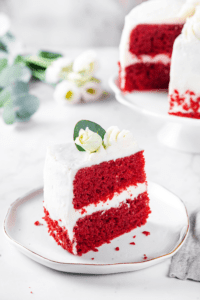 A white plate with a piece of red velvet cake on it. Part of the cake whole cake is in the background.