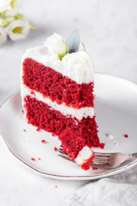 A piece of red velvet cake on a white plate. A fork is in front of the cake with a piece of cake on it.